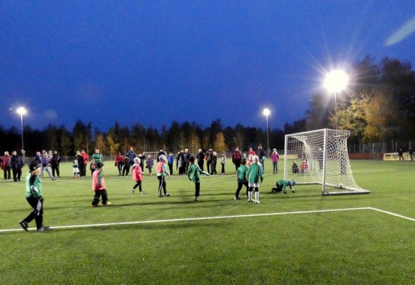 Iltapeli tekonurmikentällä lamppujen loisteessa, jossa pienet lapset pelaavat jalkapalloa.